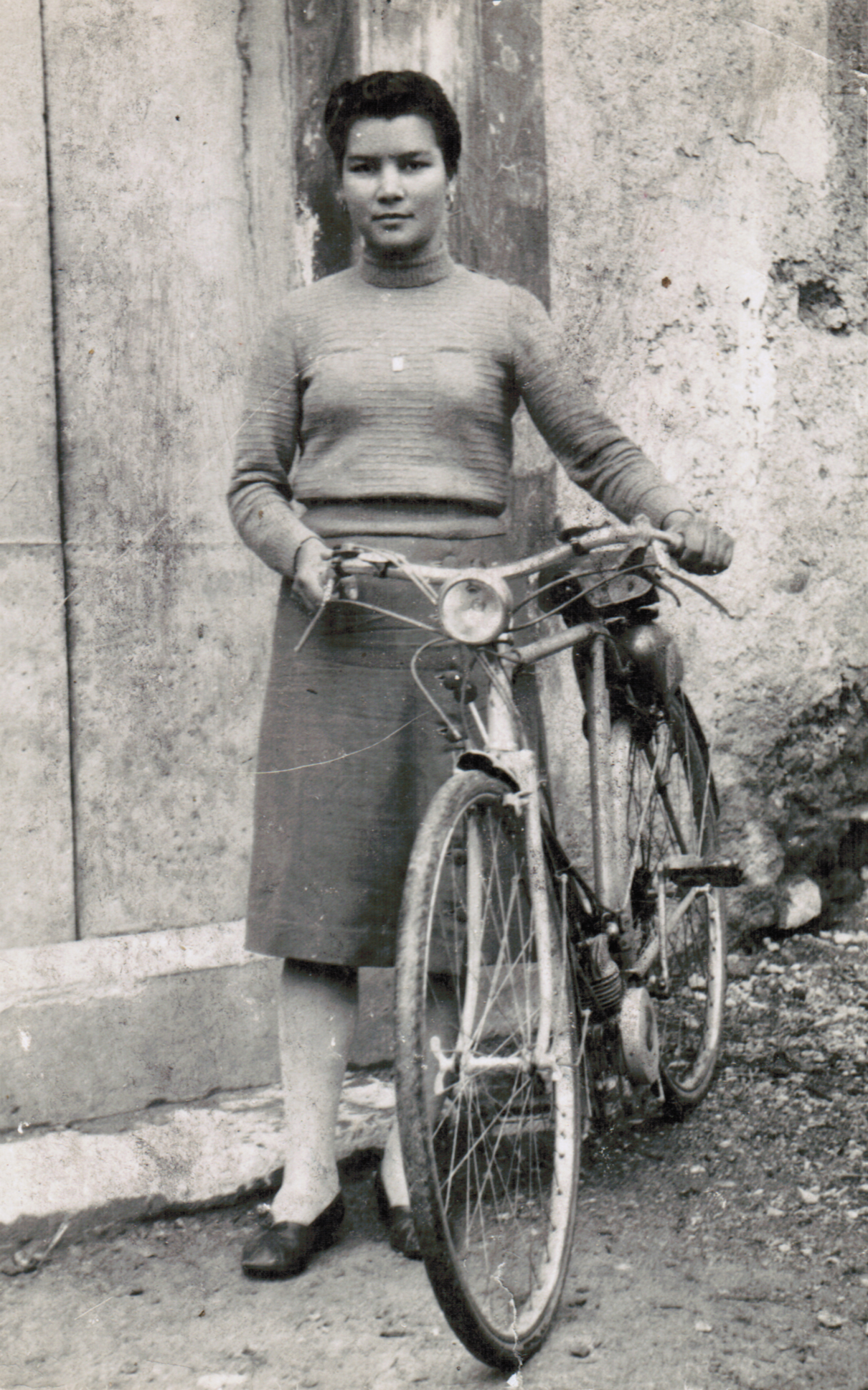 A greyscale picture of a women with a bicycle in front of a concrete wall